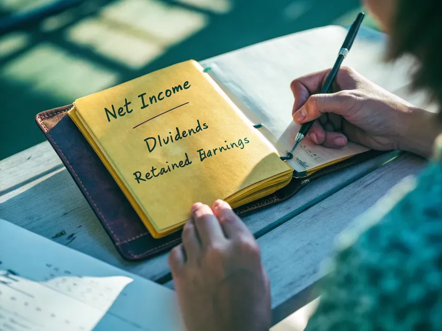 Close-up of a vintage ledger open to financial terms 'Net Income,' 'Dividends,' and 'Retained Earnings' with hands writing.